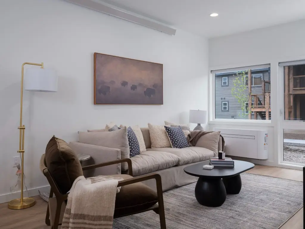 Staged one-bedroom model living room with sofa, chair, and a window onto the courtyard